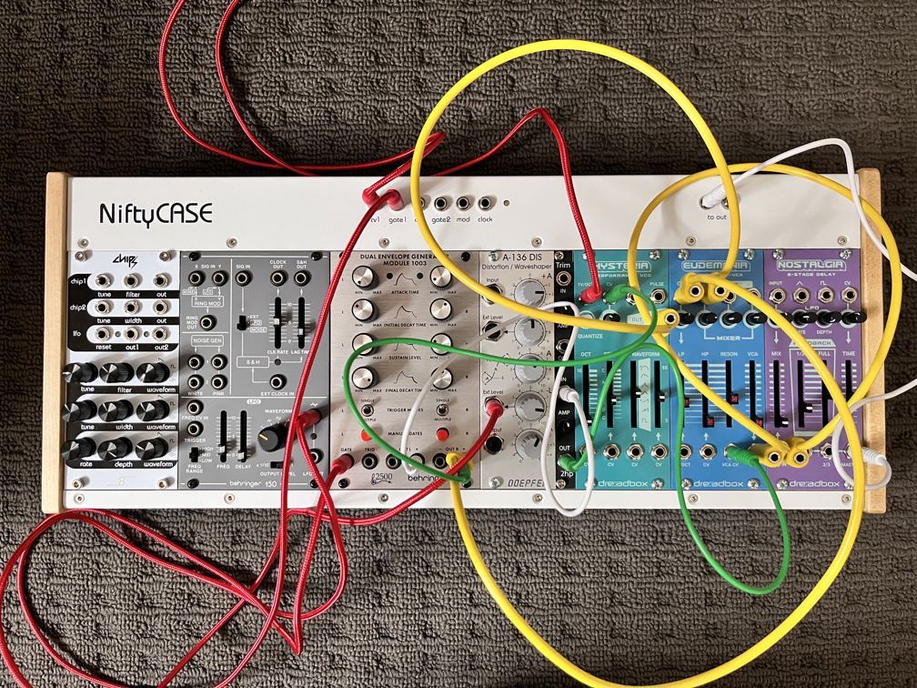 A modular synth case with yellow, red and green patch cables. The modules on the left side are different shades of grey, the ones on the right are coloured cyan, blue and purple. The case has a label "NiftyCase" in a retro-modernist typeface