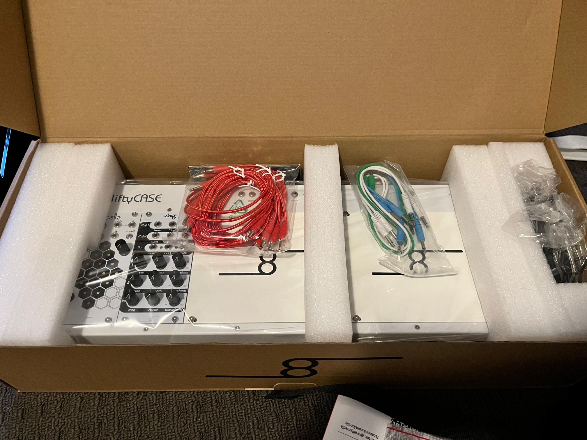 A photo of a white modular synth case in its packaging with some packs of colourful patch cables on top